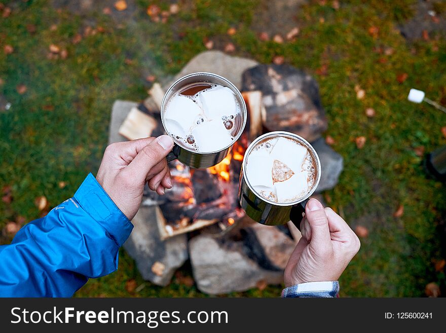 Wanderlust and Travel concept. Metal cups with hot chocolate and marshmallows. Fire on background. Atmospheric moment.
