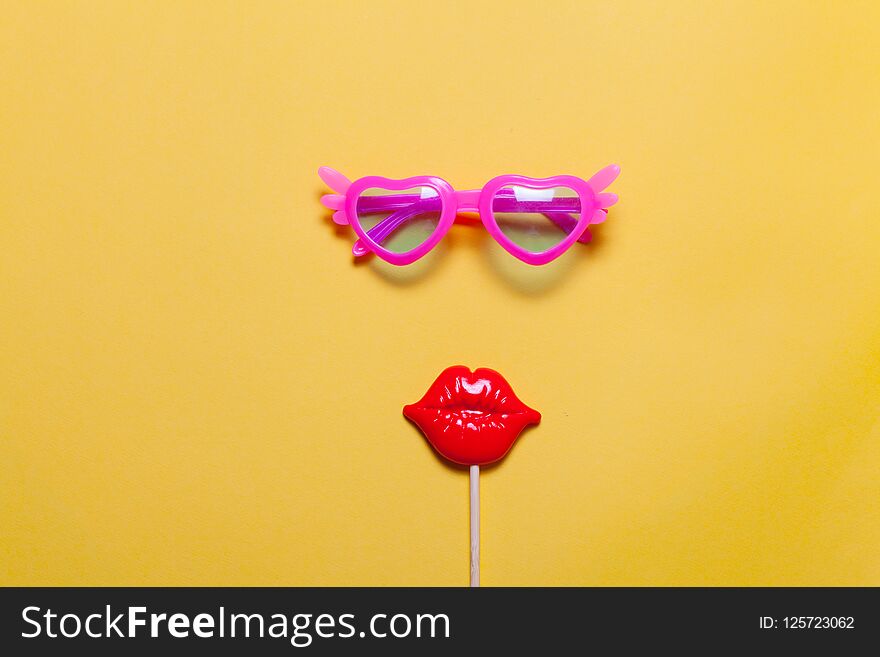 Holiday minimalistic flat lay: funny pink sunglasses and masquerade plastic lips on stick. Yellow background