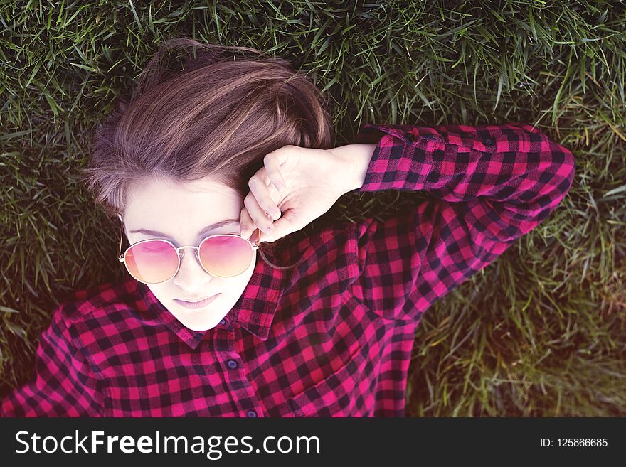 Sweet teenage girl laying on grass in park in trendy red glasses and posing for camera. Casual young woman on green grass. Sweet teenage girl laying on grass in park in trendy red glasses and posing for camera. Casual young woman on green grass.