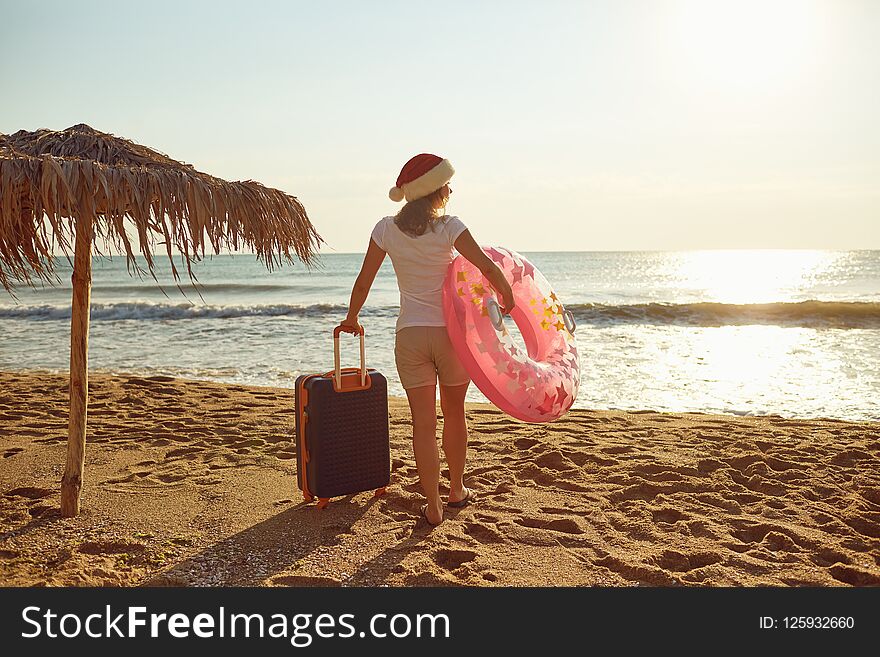 A girl in a Santa Claus hat with a suitcase on the beach on Christmas Day. The concept of Christmas by the sea.