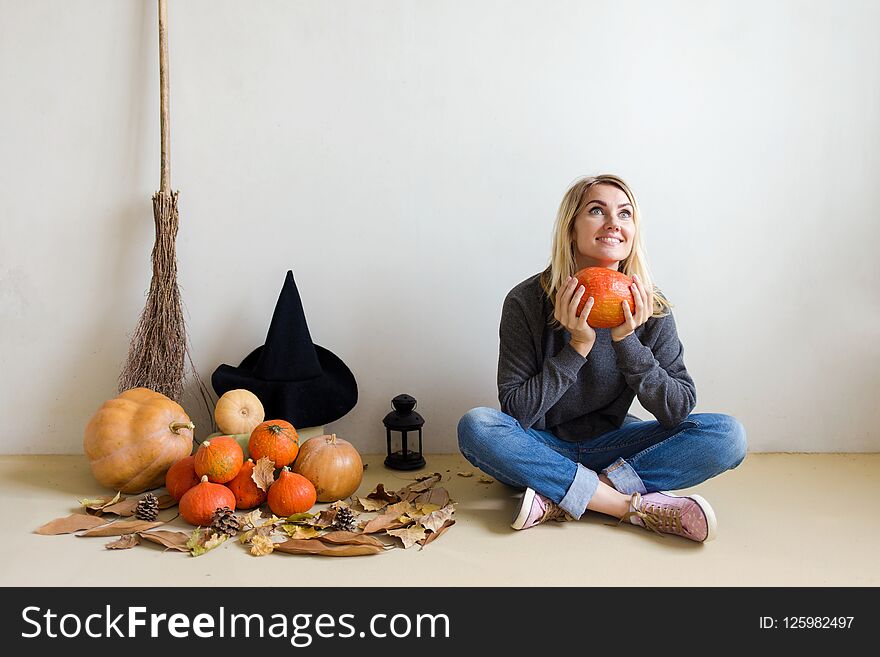 Halloween Concept. Beautiful Blond Girl With A Witch Hat With Pumpkins And A Broom Sitting On The Floor Against A White Wall Backg