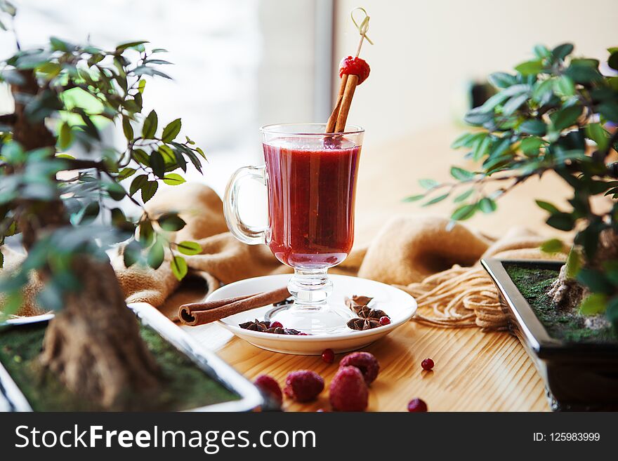 Closeup of hot mulled wine in glass with cinnamon sticks and cherry on top, standing near green plant in pot, with anise star on plate and rough cloth as decor