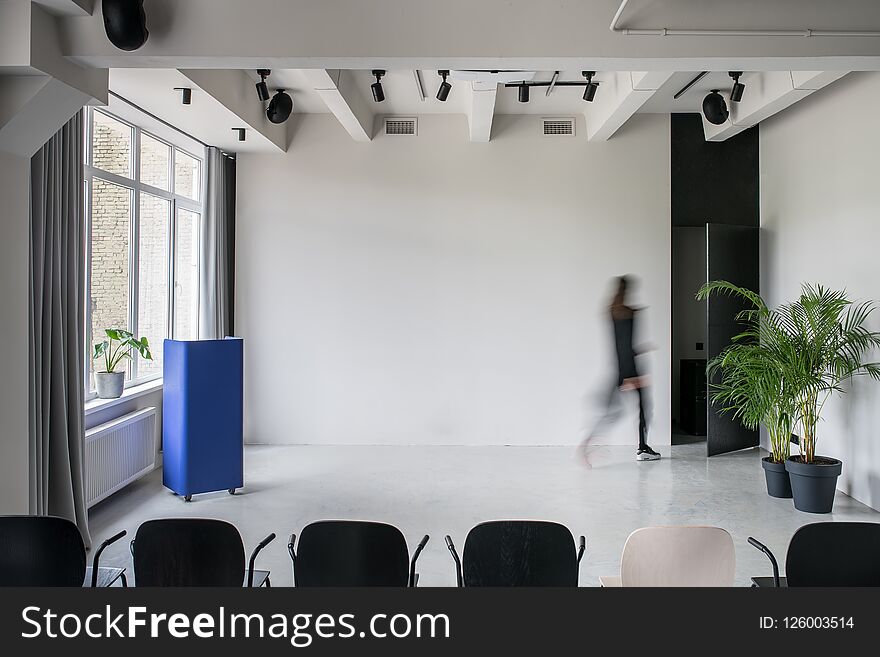 Nice conference hall with gray walls and a glossy floor. There is a row of multicolor chairs, metallic blue stand, walking person, opened door, green plants, window with curtains, lamps and speakers. Nice conference hall with gray walls and a glossy floor. There is a row of multicolor chairs, metallic blue stand, walking person, opened door, green plants, window with curtains, lamps and speakers.