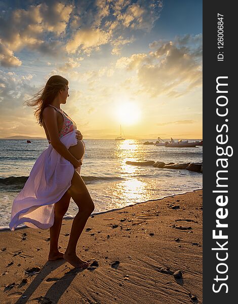 Pregnant woman in white dress and exposed belly stands at a beach during sunset time. Pregnant woman in white dress and exposed belly stands at a beach during sunset time