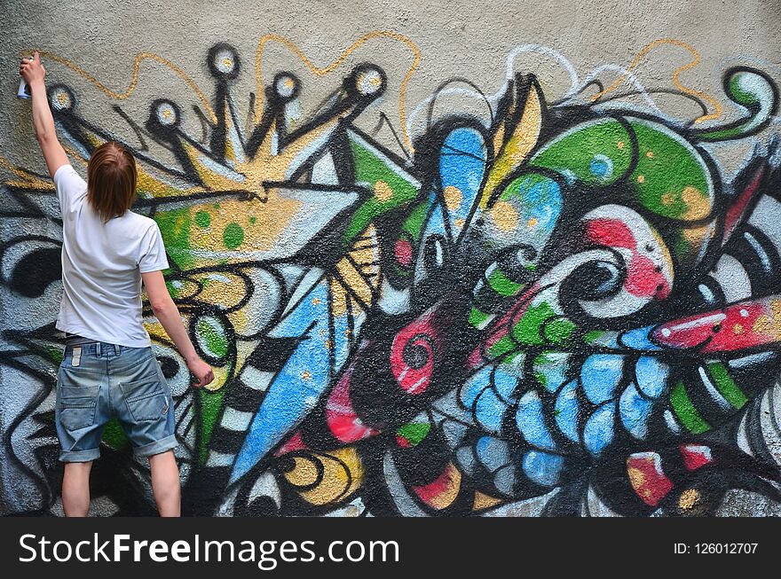 Photo of a young guy in denim shorts and a white shirt. The guy draws on the graffiti wall a drawing with aerosol paints of various colors. The concept of hooliganism and damage to property