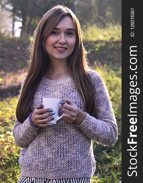 Portrait of a cheerful young smiling woman in autumn forest holding ceramic white coffee cup in autumn forest outdoors. Portrait of a cheerful young smiling woman in autumn forest holding ceramic white coffee cup in autumn forest outdoors