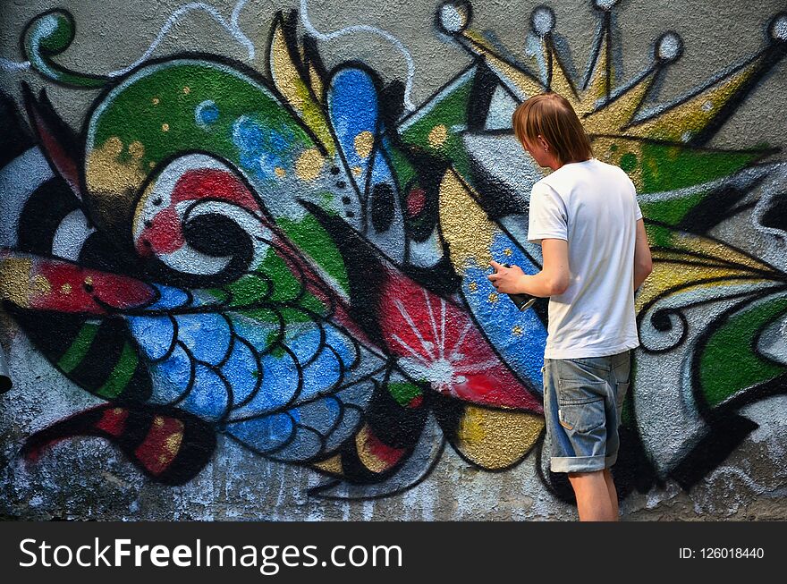 Photo of a young guy in denim shorts and a white shirt. The guy draws on the graffiti wall a drawing with aerosol paints of various colors. The concept of hooliganism and damage to property