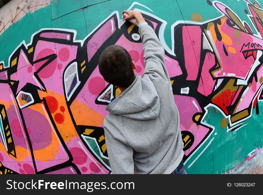 A young guy in a gray hoodie paints graffiti in pink and green colors on a wall in rainy weather. Fisheye shot