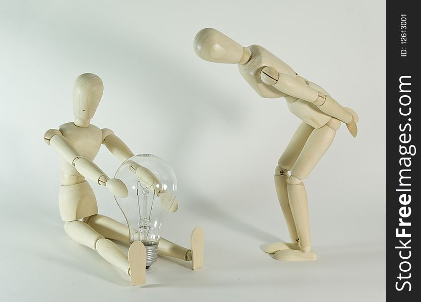 A wooden toy figure holds a light bulb while another figure watches nearby. White background. A wooden toy figure holds a light bulb while another figure watches nearby. White background