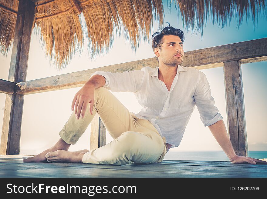 Beautiful young relaxed man in a small wooden deck. Strong summer warm lights. Trendy haircut. White shirt and beige trousers.
