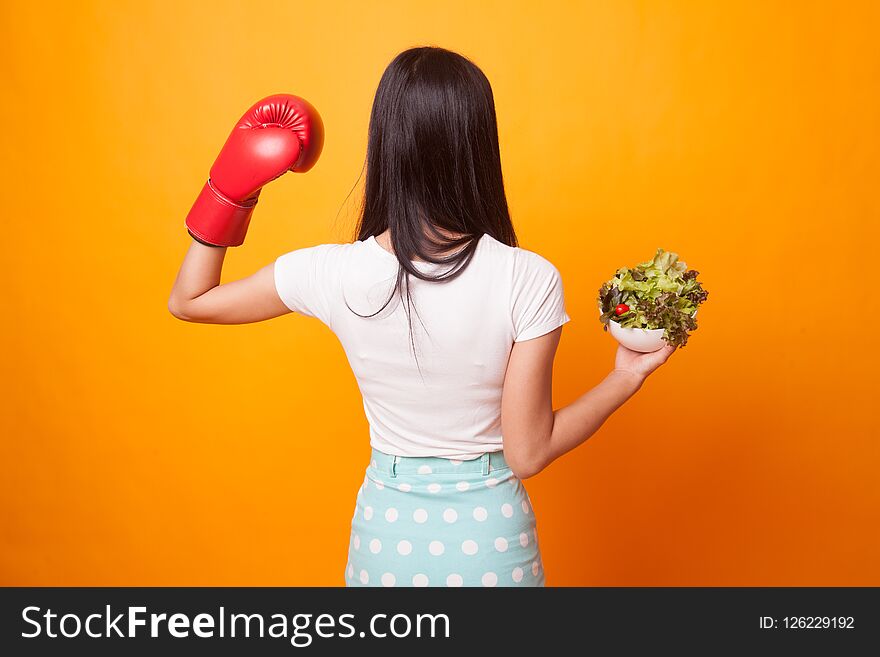 Young Asian woman with boxing glove and salad on bright yellow background. Young Asian woman with boxing glove and salad on bright yellow background