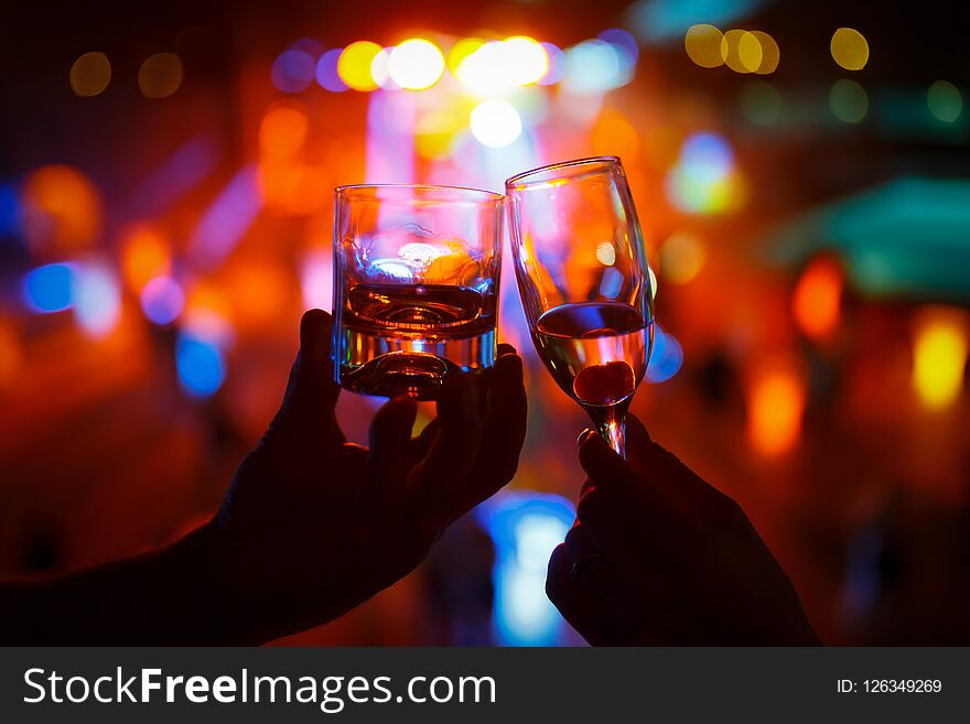 Wineglass of champagne in woman hand and a glass of whiskey in a man hand against a background of colored lights