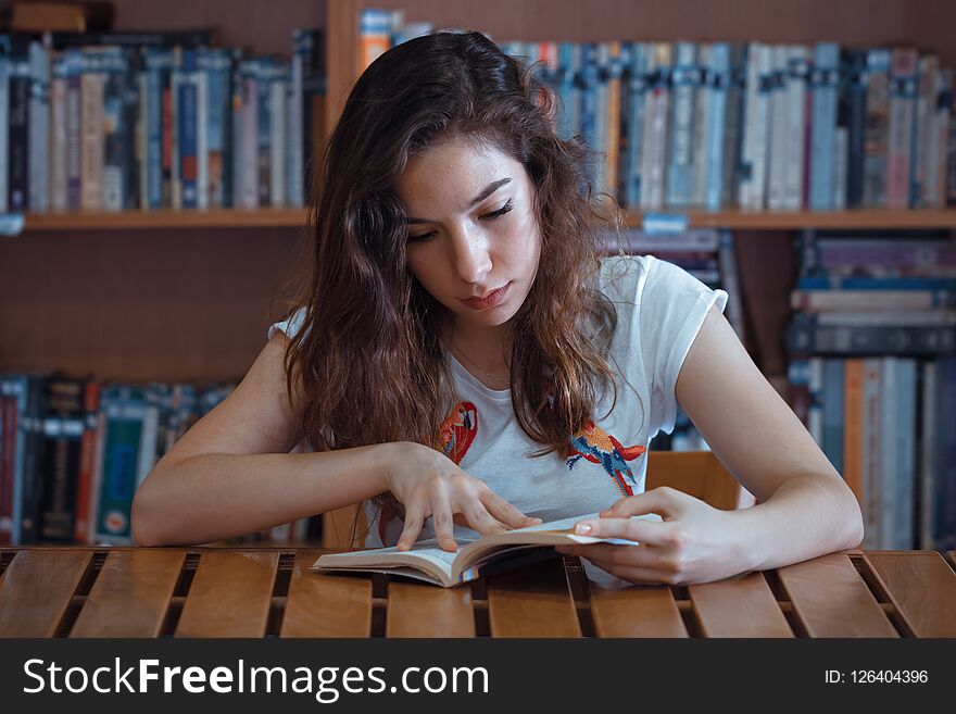 Serious young cute girl reading book in library at desk