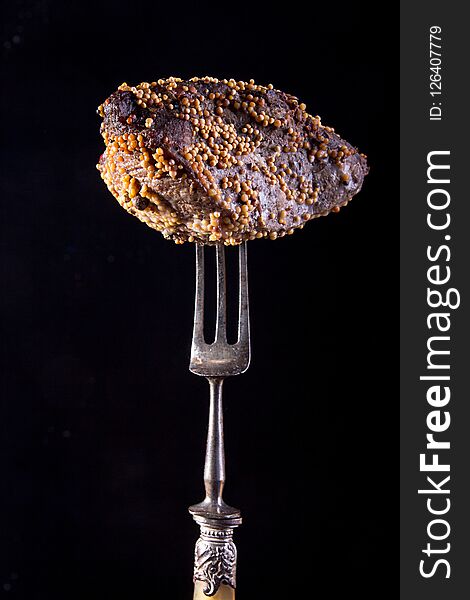 Piece of roasted meat on fork. Cold-boiled baked pork with mustard grains on black background.