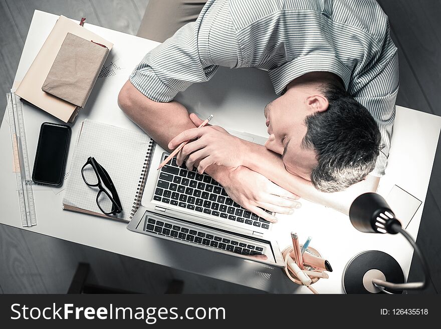 Feeling exhausted. Top view of handsome office worker sleeping on the table holding pencil in hand. Feeling exhausted. Top view of handsome office worker sleeping on the table holding pencil in hand