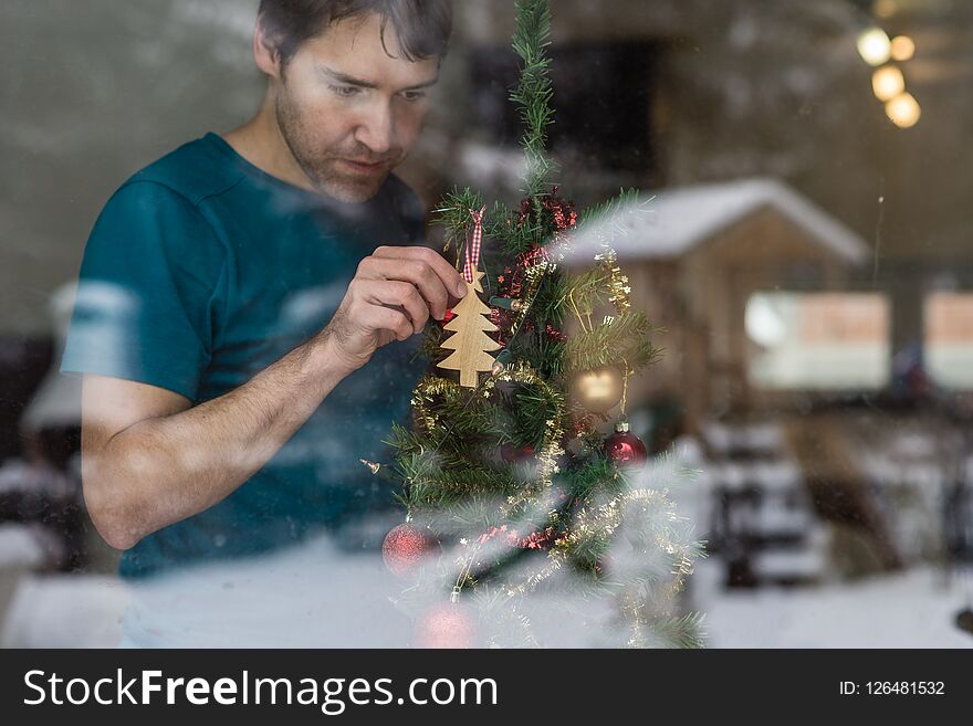 View through a window of a young man hanging wooden tree on a Christmas tree with reflection of winter nature in glass.