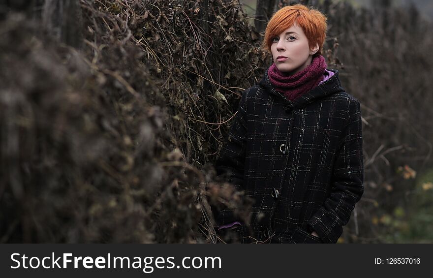 Sweet red-haired girl in a black coat and purple knitted scarf is standing by the fence overgrown with grapevine or ivy, girl or w