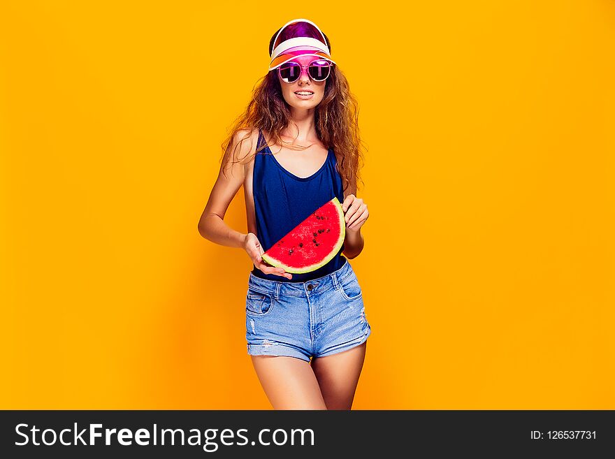 Beautiful young female in cap, sunglasses and swimsuit hold slice of watermelon and looking away while standing on yellow background. Copyspase. Beautiful young female in cap, sunglasses and swimsuit hold slice of watermelon and looking away while standing on yellow background. Copyspase