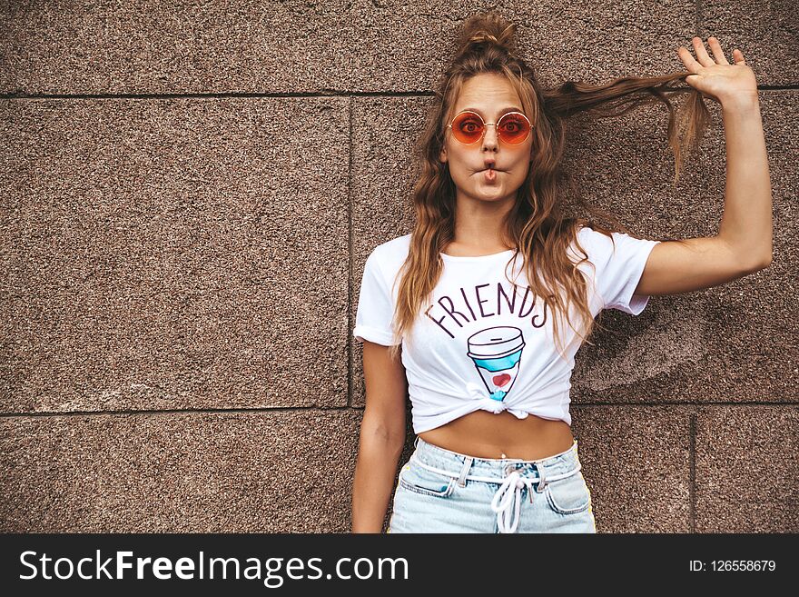 Beautiful cute blond teenager model in summer hipster white clothes posing near wall on the street. Making funny fish face. Beautiful cute blond teenager model in summer hipster white clothes posing near wall on the street. Making funny fish face