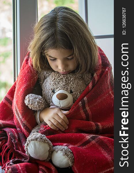 Sad little girl sitting on the windowsill hiding behind an old checkered plaid and carefully embracing her beloved toy bear. Sad little girl sitting on the windowsill hiding behind an old checkered plaid and carefully embracing her beloved toy bear