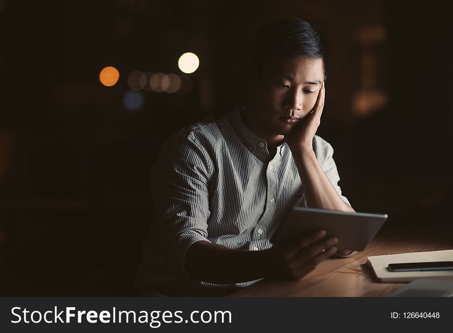 Tired young Asian businessman using a digital tablet while sitting in an office late at night with city lights glowing in the background. Tired young Asian businessman using a digital tablet while sitting in an office late at night with city lights glowing in the background