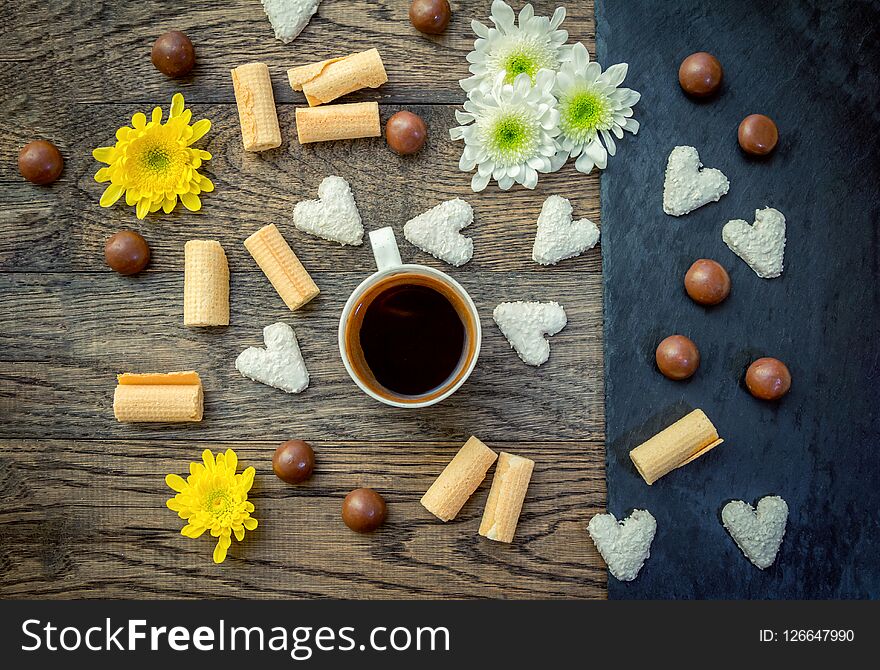 Coffee in a cup, sweets and flowers on the table – breakfast or snack. Chocolate candies, heart shape cookies, waffle rolls, white and yellow flowers on a wooden table.