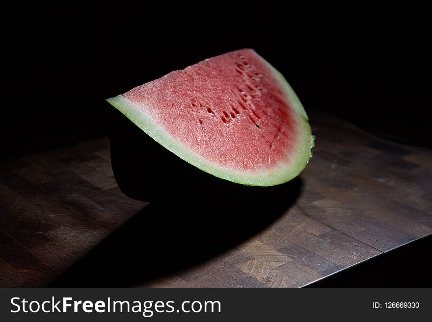 Watermelon slice on chopping board extreme closeup on black background with harsh light. Watermelon slice on chopping board extreme closeup on black background with harsh light