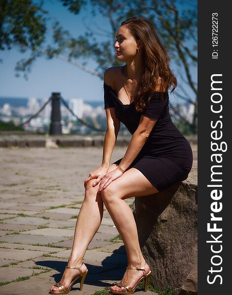 Portrait of a beautiful girl in a stylish dress sitting on a stone on a background of a cityscape