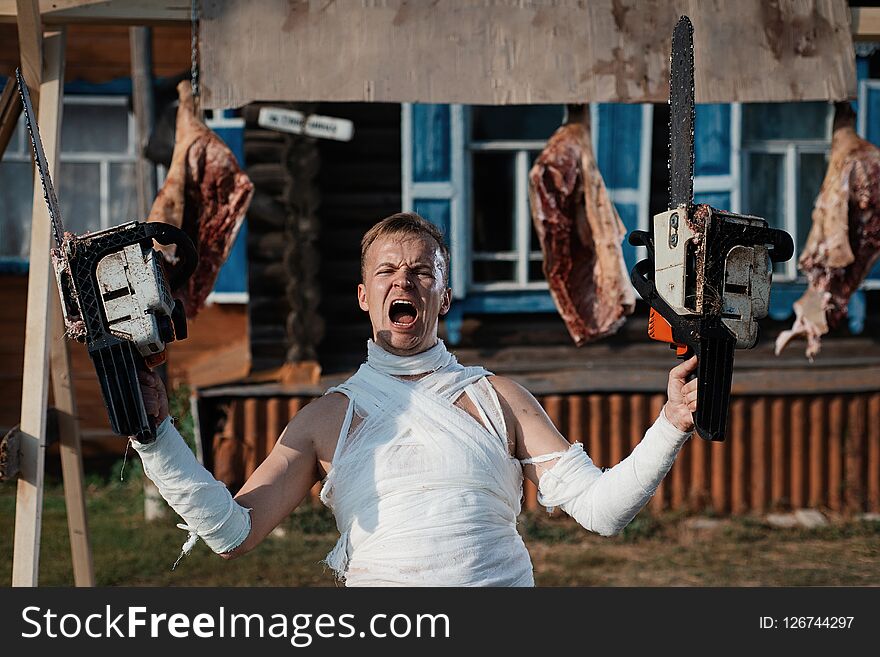 Bandaged man screams terribly, holding two chainsaws in his hands on background of pig carcasses