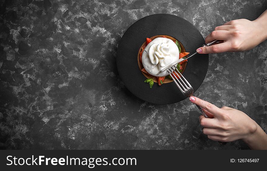 Woman eating burrata cheese on small wooden plate served with fresh tomatoes and basil on dark textured background. Studio shot