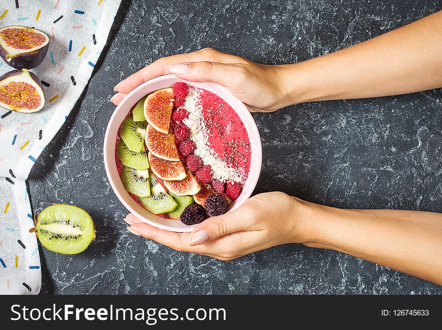 Woman hands hold raspberries smoothie bowl with figs, kiwi and coconut on concrete background. Top view