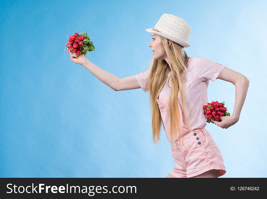 Positive teenage blonde long hair girl wearing summer clothing sun hat holding two bunches of fresh radish, on blue. Positive teenage blonde long hair girl wearing summer clothing sun hat holding two bunches of fresh radish, on blue