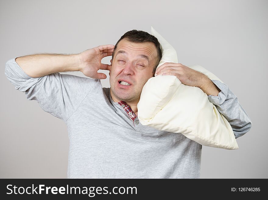 Sleepy tired angry guy holding pillow in one and closing his ear with another hand protecting from noise, on grey. Health care, sleep disorder concept. Sleepy tired angry guy holding pillow in one and closing his ear with another hand protecting from noise, on grey. Health care, sleep disorder concept