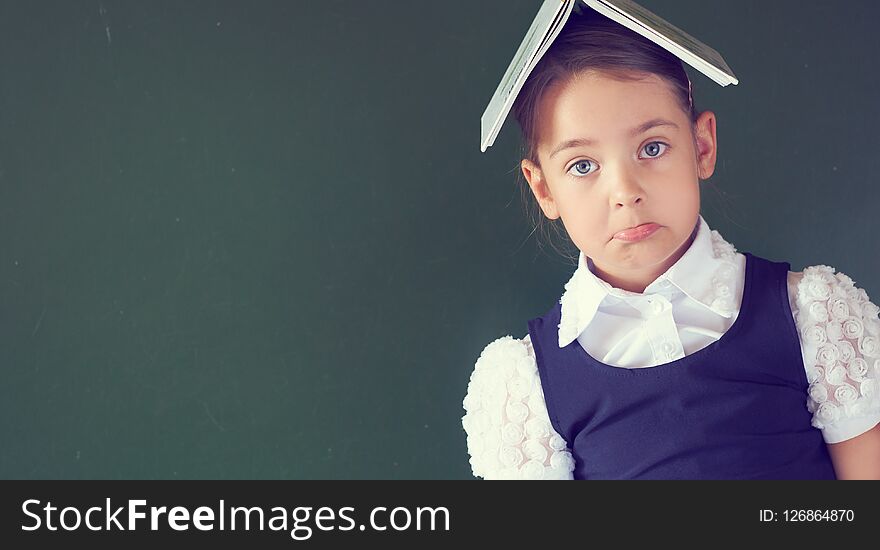 Pretty schoolgirl in a school dress stands holding an open book on her head near at the blackboard. Little girl does not know the answer to the teacher`s question