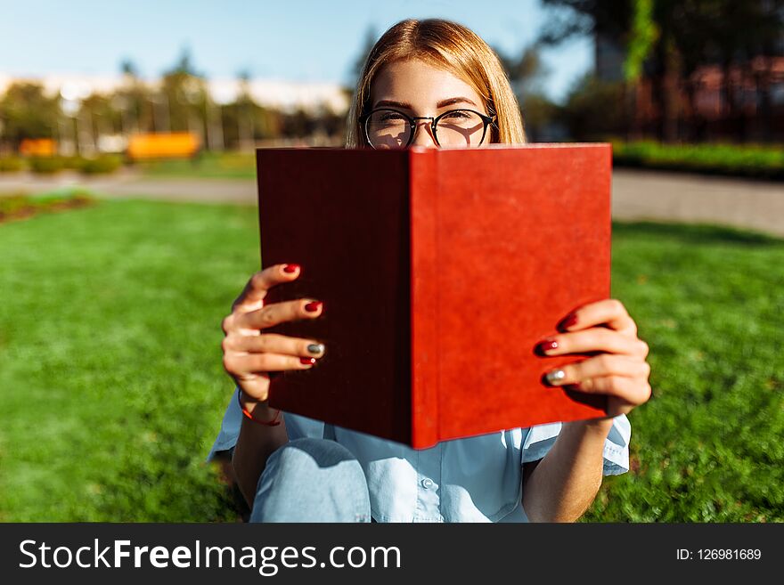 Portrait of cheerful student girl sitting outdoors on green grass, wearing glasses, holding book near her face. Portrait of cheerful student girl sitting outdoors on green grass, wearing glasses, holding book near her face