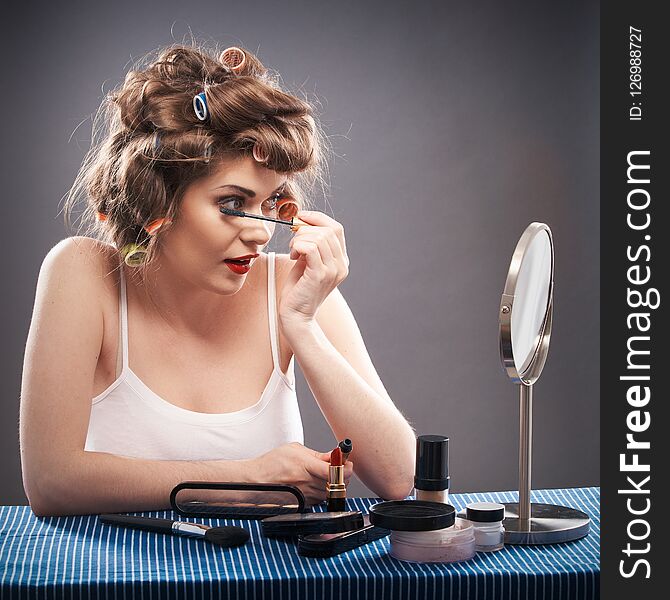 Portrait of a young woman with long hair on gray background making beauty face and hair style. Smile happy girl seating at table with make up accessories and mirror, creating home beauty salon. Portrait of a young woman with long hair on gray background making beauty face and hair style. Smile happy girl seating at table with make up accessories and mirror, creating home beauty salon