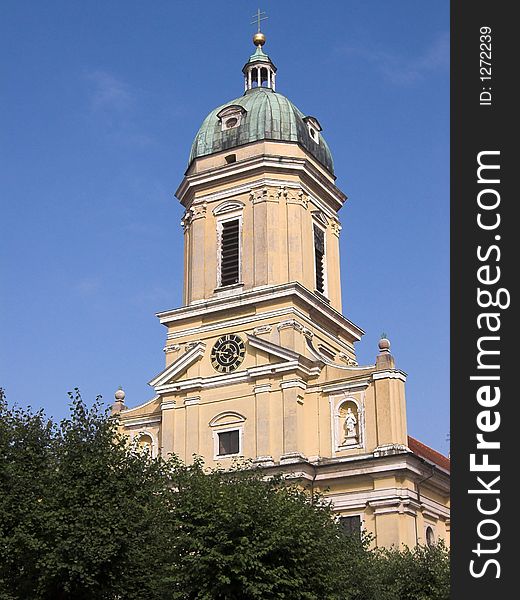 The image shows a church tower in the city of Neuburg (Bavaria, Germany. In the background of the image is a blue sky, in front of the chapel are some green bushes.