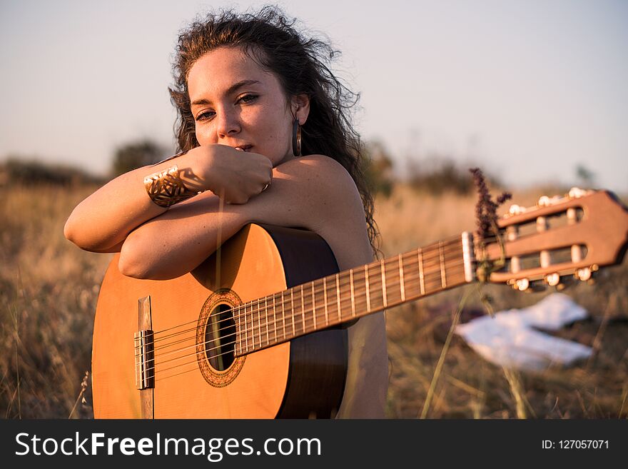 Naked Contemplative Curly brunette girl Sitting with guitar in the field with taked off clothes on background