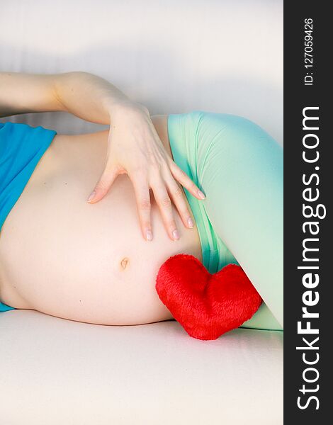 Pregnant woman future mother lying on couch with red heart love symbol, caressing her naked belly. Pregnancy, motherhood and baby anticipation concept. Pregnant woman future mother lying on couch with red heart love symbol, caressing her naked belly. Pregnancy, motherhood and baby anticipation concept.