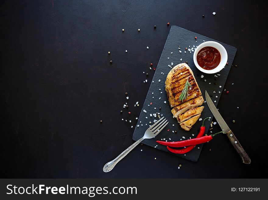 Grilled chicken fillets on slate plate with rosemary, pepper, ketchup and spices on dark wooden background. Top view. Flat lay. Copy space