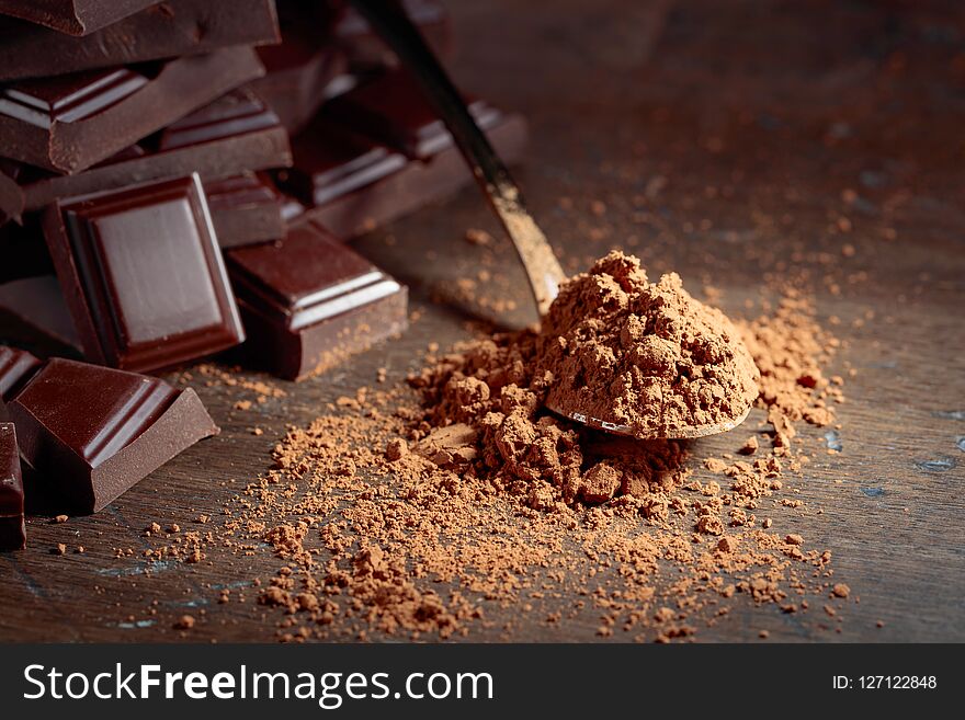 Broken chocolate pieces and cocoa powder in small spoon on a wooden background. Copy space. Broken chocolate pieces and cocoa powder in small spoon on a wooden background. Copy space.
