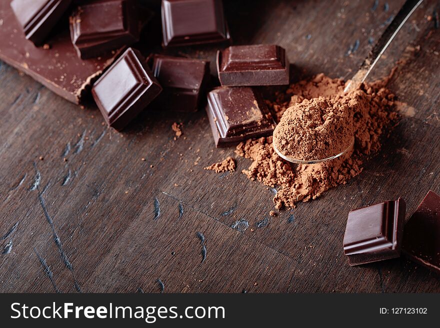 Broken chocolate pieces and cocoa powder in small spoon on a wooden background. Copy space. Broken chocolate pieces and cocoa powder in small spoon on a wooden background. Copy space.