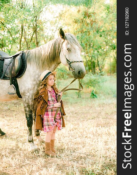 A nice little girl with light curly hair in a vintage plaid dress and a straw hat and a gray horse. Rural life in autumn. Horses and people