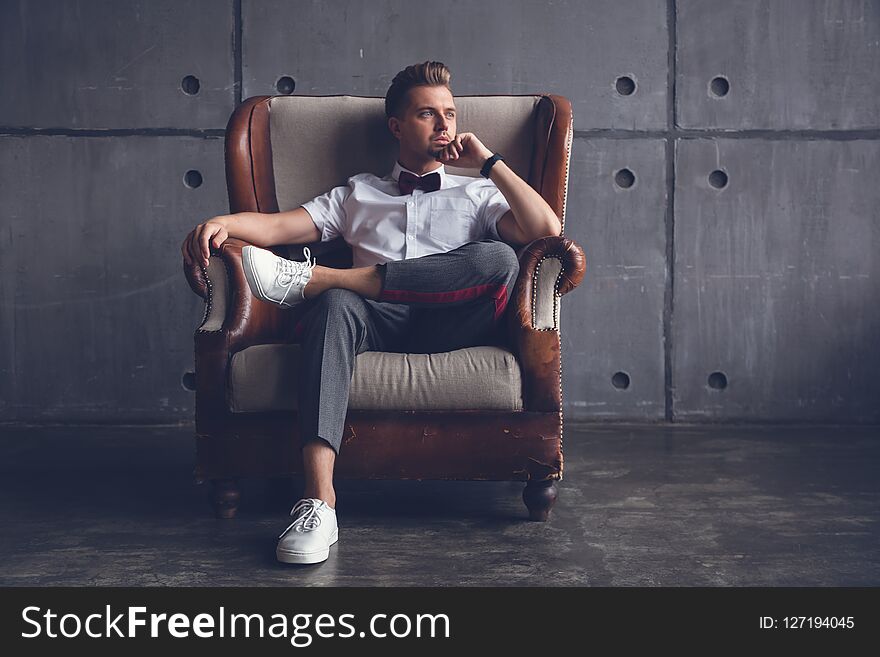 Young attractive serious glamorous male hipster in white shirt and butterfly sitting in chair, on gray background