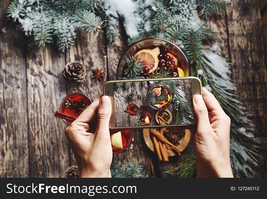 Woman taking pictures of of glass bowl with Christmas mulled wine and ingredients on rustic wooden background with spruce branches using phone. Woman Food photographer with hands visible photographing for the social networks. Woman taking pictures of of glass bowl with Christmas mulled wine and ingredients on rustic wooden background with spruce branches using phone. Woman Food photographer with hands visible photographing for the social networks.