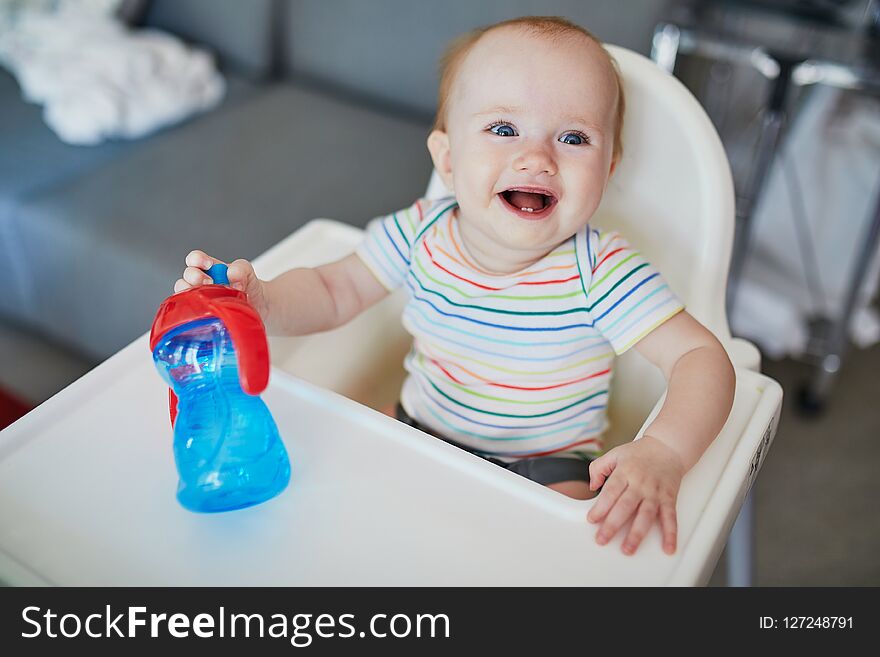 Little baby girl sitting in high chair at home or at restaurant and drinking water from sippy cup