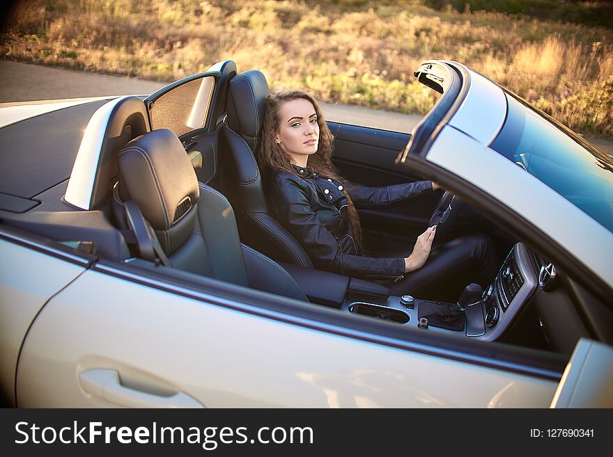 Beautiful girl with long hair in a leather jacket and leather pants in sunglasses sits in an expensive car at sunset.