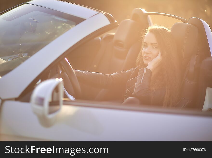 Beautiful girl with long hair in a leather jacket and leather pants in sunglasses sits in an expensive car at sunset.