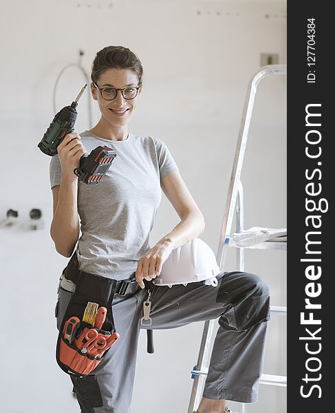 Professional repairwoman with tool belt and safety helmet doing a home renovation, she is posing and holding a drill. Professional repairwoman with tool belt and safety helmet doing a home renovation, she is posing and holding a drill