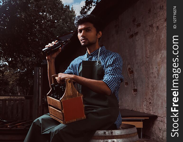 Young Indian brewer in uniform sitting on a wooden barrel and drink craft beer.